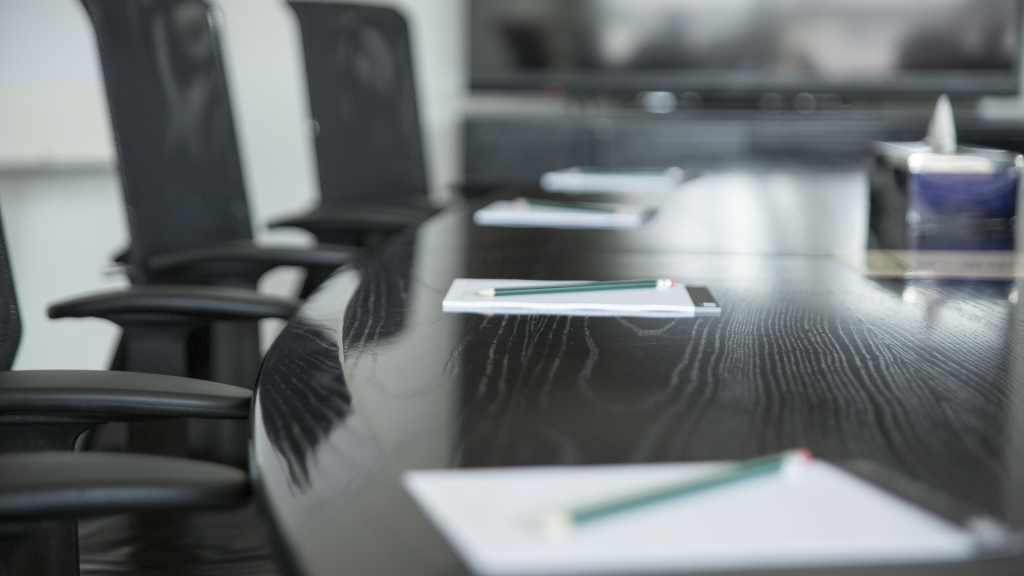 Mesa de reuniones circular con documentos y lápices preparados, representando el proceso de planificación y estrategia en una adquisición empresarial, con una pantalla de fondo para presentaciones.