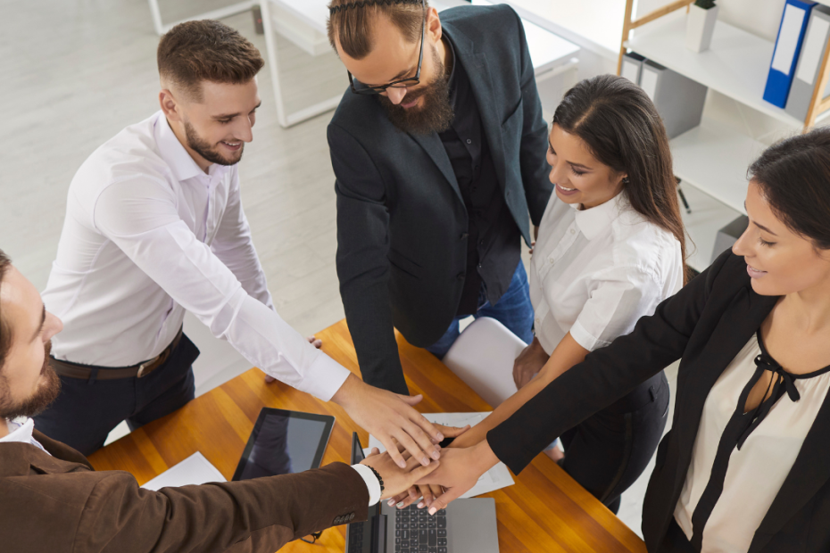 A team of professionals joining hands in a symbol of collaboration, representing the collaborative work of a Temporary Business Association (UTE), with documents and technology on the table in an office setting.