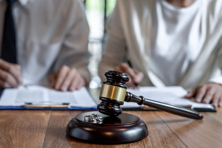 Dos personas firmando diferentes documentos, con un mazo de juez en primer plano y dos anillos encima de él, haciendo referencia al procedimiento de divorcio.