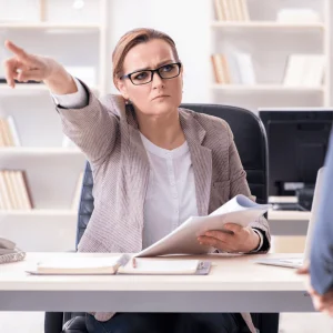 Mujer seria con gafas y ropa de oficina, sentada en su escritorio y señalando hacia la puerta mientras sostiene unos documentos, en actitud de despedir a un trabajador.