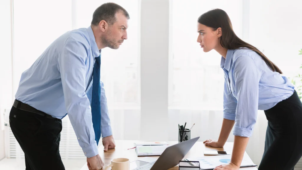 Un hombre y una mujer, ambos vestidos con ropa formal de oficina, están enfrentados cara a cara sobre una mesa de reuniones, en una postura corporal que refleja tensión y desacuerdo. Están inclinados hacia adelante con las manos apoyadas en la mesa, mirándose fijamente, lo que sugiere un conflicto o discusión intensa. En la mesa hay documentos, una computadora portátil y una taza. La escena se desarrolla en una oficina luminosa con ventanas grandes. La imagen representa un conflicto laboral, ideal para ilustrar temas como mecanismos legales para resolver conflictos entre socios.