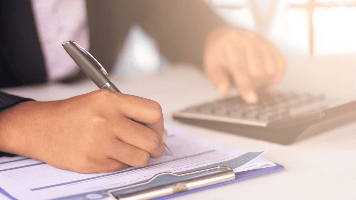 Primer plano de una persona con traje escribiendo con bolígrafo sobre un formulario sujetado por una carpeta azul, mientras usa una calculadora con la otra mano. La imagen transmite una escena de trabajo administrativo o contable, en un ambiente bien iluminado. La acción sugiere revisión o registro de datos financieros.
