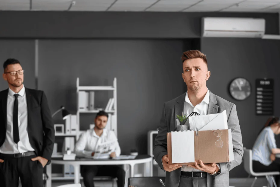Un hombre joven, vestido con traje gris y expresión seria, sostiene una caja de cartón con objetos personales como una planta, carpetas y unas gafas, simbolizando su salida de la empresa. Detrás de él, en una oficina moderna con paredes grises, se observan otros empleados trabajando y un hombre con traje oscuro que lo mira desde la distancia. La escena representa la expulsión de un socio o empleado del entorno laboral.