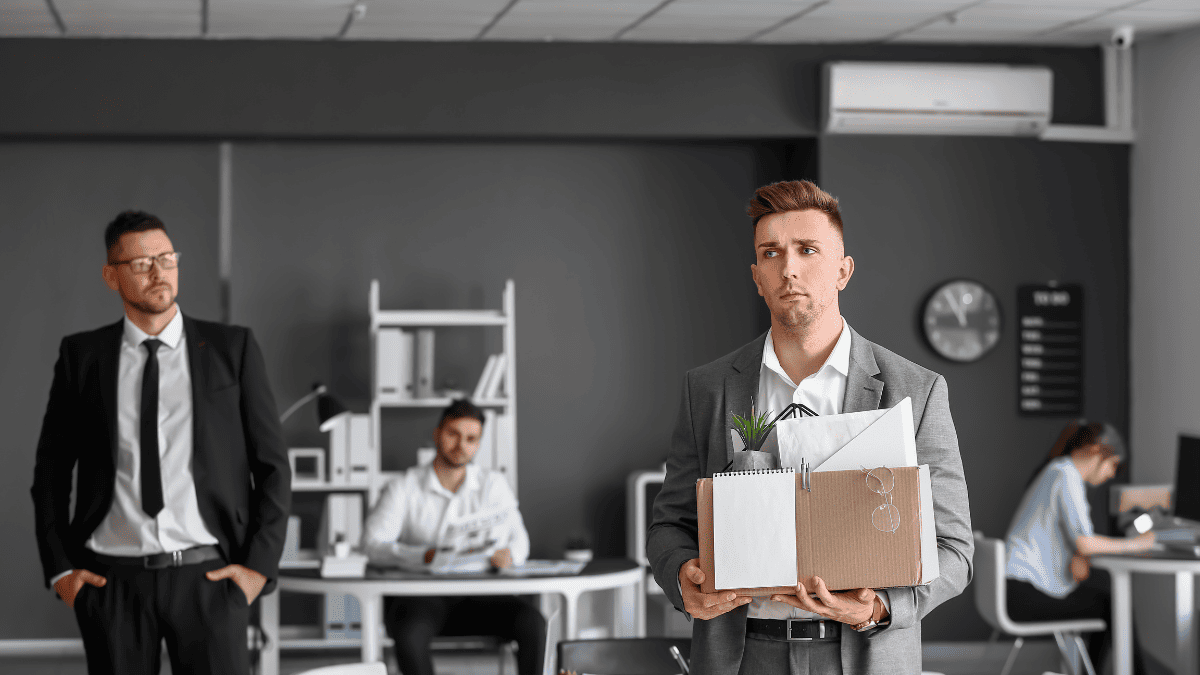 Un hombre joven, vestido con traje gris y expresión seria, sostiene una caja de cartón con objetos personales como una planta, carpetas y unas gafas, simbolizando su salida de la empresa. Detrás de él, en una oficina moderna con paredes grises, se observan otros empleados trabajando y un hombre con traje oscuro que lo mira desde la distancia. La escena representa la expulsión de un socio o empleado del entorno laboral.