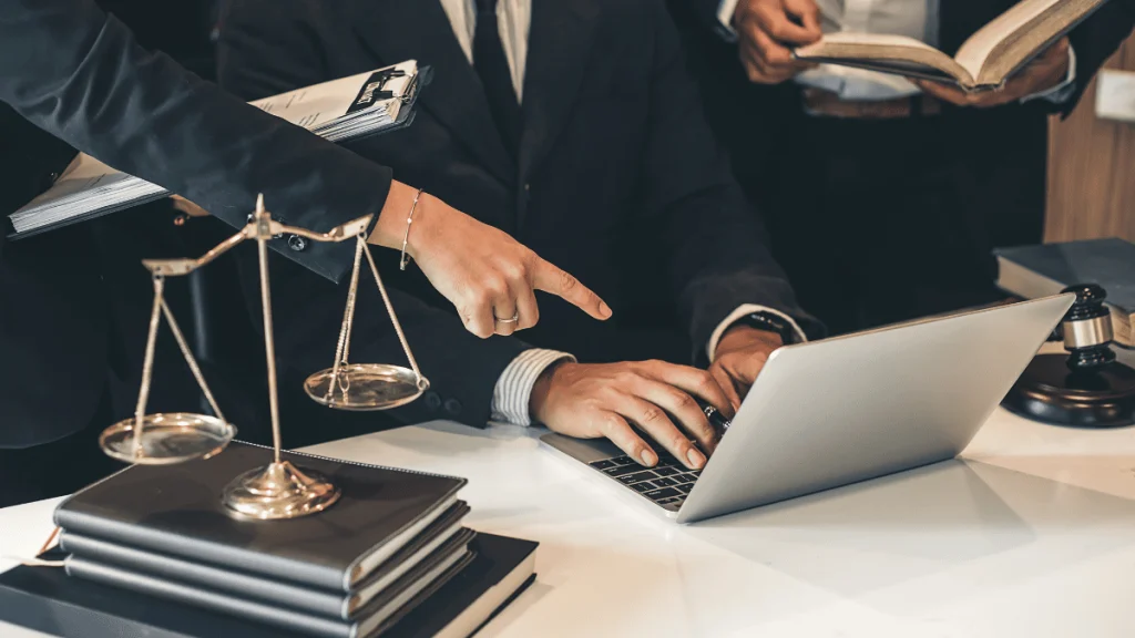 Tres personas con vestimenta formal están reunidas en una oficina legal. En primer plano, una mano señala la pantalla de una computadora portátil mientras otra persona escribe en el teclado. Sobre el escritorio hay una balanza de la justicia metálica, una pila de libros y una carpeta, símbolos representativos del ámbito jurídico. Al fondo, una tercera persona sostiene un libro abierto. La escena transmite un ambiente profesional y colaborativo, típico de un despacho de abogados. La imagen evoca el concepto de asesoría legal.