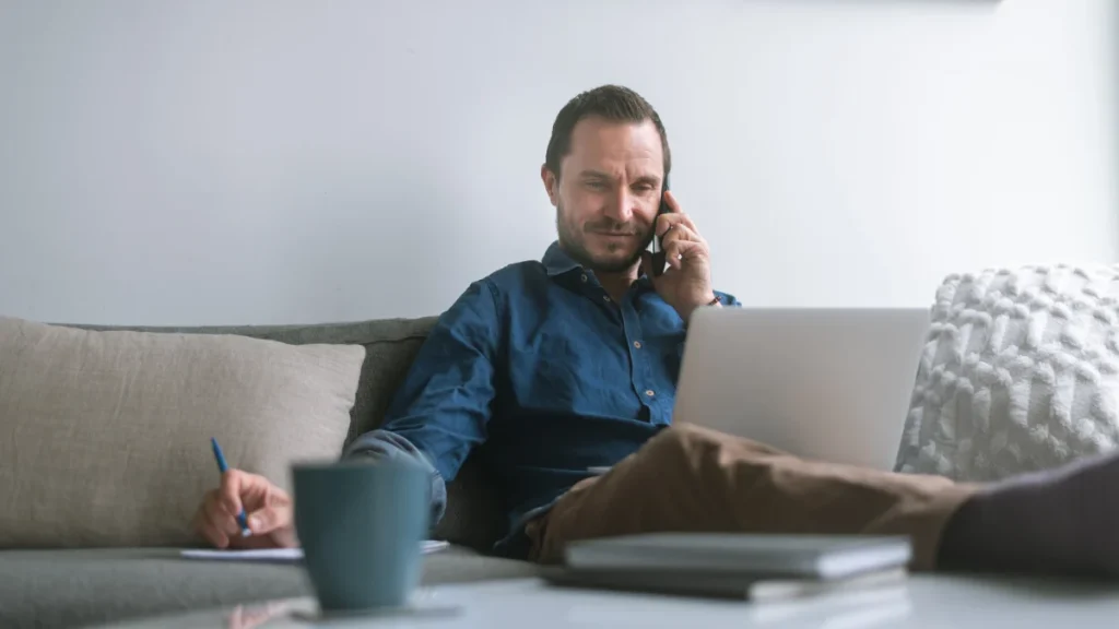 Hombre teletrabajando desde casa con portátil y teléfono móvil mientras revisa contratos laborales internacionales