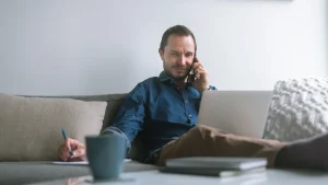 Hombre teletrabajando desde casa con portátil y teléfono móvil mientras revisa contratos laborales internacionales