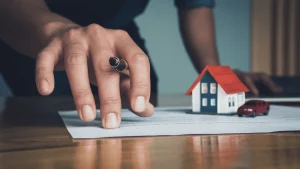 Primer plano de una mano masculina sosteniendo una pluma sobre un documento legal, en una escena que sugiere la firma o revisión de un contrato. Junto al papel, sobre la mesa, hay una maqueta de casa con techo rojo y una figura de automóvil rojo, simbolizando bienes como propiedades y vehículos. Al fondo, otra mano se apoya sobre una computadora portátil, indicando un entorno administrativo o legal. La imagen representa el concepto de bienes materiales en el contexto de procesos jurídicos, adecuada para ilustrar temas como la defensa legal en casos de alzamiento de bienes.