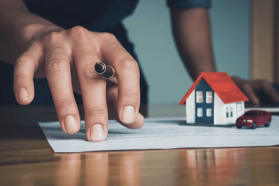 Primer plano de una mano masculina sosteniendo una pluma sobre un documento legal, en una escena que sugiere la firma o revisión de un contrato. Junto al papel, sobre la mesa, hay una maqueta de casa con techo rojo y una figura de automóvil rojo, simbolizando bienes como propiedades y vehículos. Al fondo, otra mano se apoya sobre una computadora portátil, indicando un entorno administrativo o legal. La imagen representa el concepto de bienes materiales en el contexto de procesos jurídicos, adecuada para ilustrar temas como la defensa legal en casos de alzamiento de bienes.