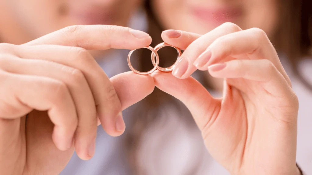 Primer plano de las manos de una pareja sosteniendo dos anillos de boda dorados, uno en cada mano, haciendo que se toquen en el centro de la imagen. Al fondo, desenfocado, se distinguen sus rostros parcialmente sonrientes, transmitiendo un momento de amor y compromiso. La imagen simboliza el enlace matrimonial y la unión entre dos personas.