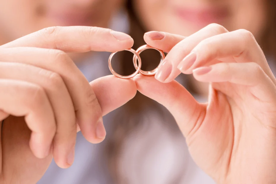 Primer plano de las manos de una pareja sosteniendo dos anillos de boda dorados, uno en cada mano, haciendo que se toquen en el centro de la imagen. Al fondo, desenfocado, se distinguen sus rostros parcialmente sonrientes, transmitiendo un momento de amor y compromiso. La imagen simboliza el enlace matrimonial y la unión entre dos personas.