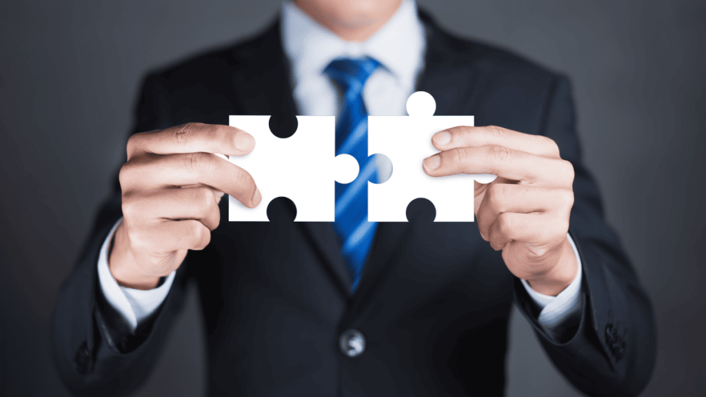 Hombre vestido con traje oscuro y corbata azul sostiene dos piezas de rompecabezas blancas frente a él, una en cada mano, alineándolas para encajarlas. La imagen representa conceptos como conexión, colaboración, solución de problemas o integración empresarial.