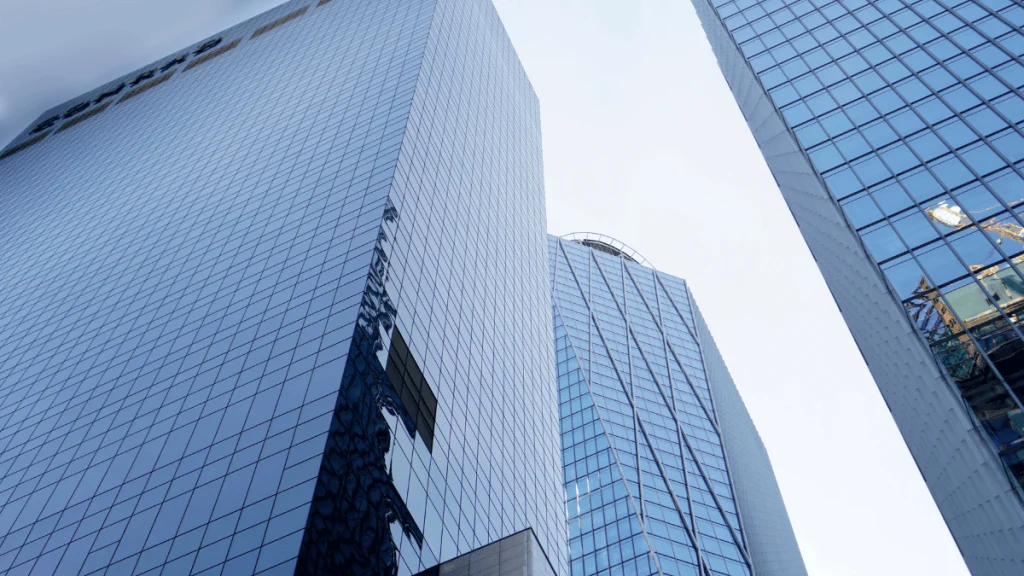 Vista en contrapicado de varios rascacielos modernos con fachadas de vidrio azul reflejante, ubicados en un distrito financiero. Las estructuras, de líneas limpias y diseño contemporáneo, simbolizan el poder corporativo y la consolidación empresarial. La imagen transmite una sensación de altura, control y estrategia, evocando el contexto de las OPAs (ofertas públicas de adquisición) y su impacto en el entorno empresarial competitivo.
