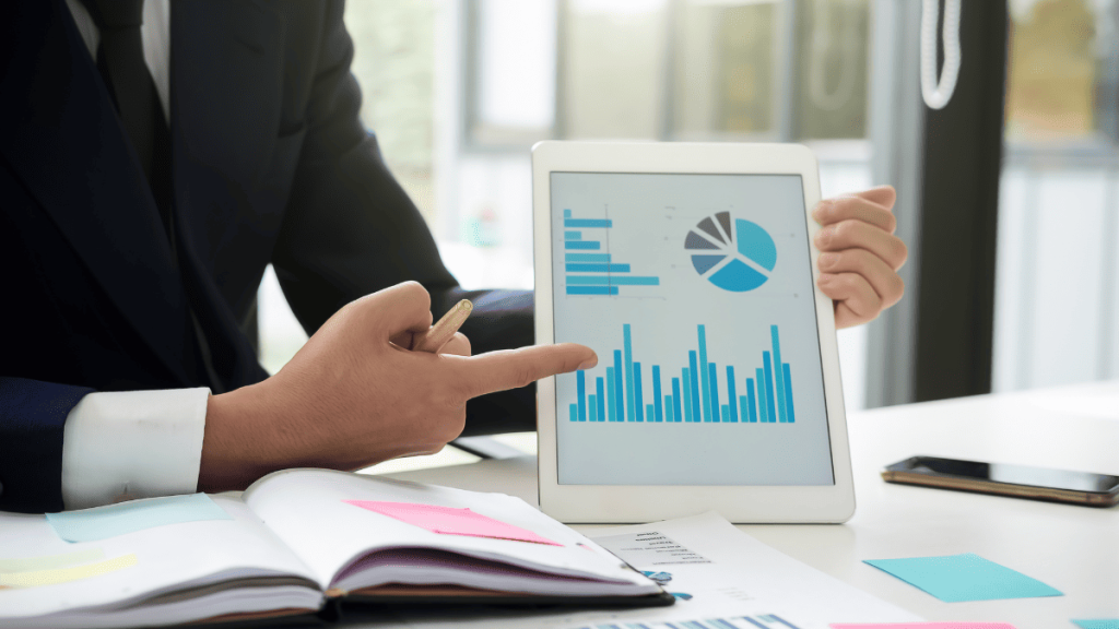 Un hombre con traje oscuro señala con un lápiz una tableta digital que muestra gráficos financieros en una oficina iluminada. En la pantalla se observan una gráfica de barras, una gráfica circular y un gráfico horizontal, todos en tonos azules. La persona parece estar explicando datos o resultados de un informe. En la mesa hay documentos, post-its de colores y un teléfono móvil. La imagen refleja un entorno de análisis de datos empresariales.