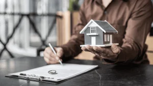Persona con camisa marrón sostiene una maqueta de casa gris con techo a dos aguas en una mano, mientras con la otra firma un contrato colocado sobre una mesa negra. El documento está sujetado por una pinza metálica y tiene un llavero con una llave. La escena representa una transacción inmobiliaria, como la compra, venta o alquiler de una vivienda.