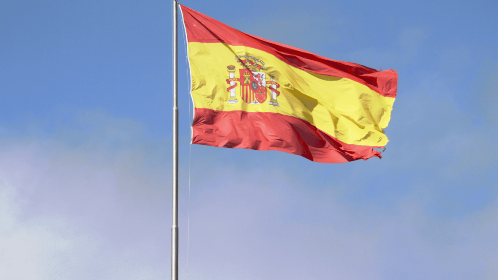 Bandera de España ondeando al viento sobre un mástil metálico, con un cielo azul claro de fondo. La bandera tiene tres franjas horizontales: roja en la parte superior e inferior, y una franja amarilla más ancha en el centro.