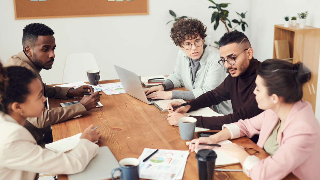 5 personas en una empresa sentadas y conversando alrededor de una mesa de color marrón, con documentos y ordenadores