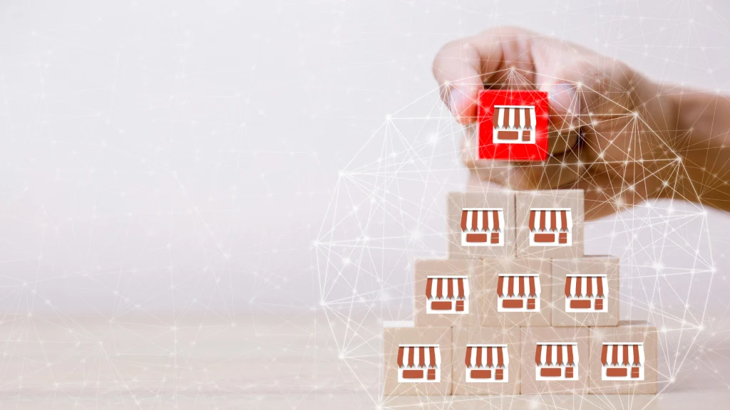 Mano sosteniendo un bloque rojo con el ícono de una tienda, colocándolo en la cima de una pirámide formada por otros bloques de madera con el mismo ícono en color blanco. La imagen sugiere una estructura jerárquica y representa el concepto de expansión de negocios bajo un contrato de franquicia internacional. El fondo muestra una red de conexiones digitales, simbolizando la globalización y la interconexión entre sucursales en distintos países.