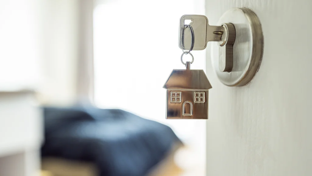 Puerta blanca de una habitación abierta, con una llave en la cerradura y un llavero con forma de casa. Al fondo de la habitación se puede una cama observar una cama con sábanas de color azul oscuro y a la izquierda un mueble blanco.