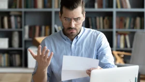 Hombre joven con gafas y camisa azul clara sentado frente a una computadora portátil, leyendo una hoja de papel con expresión de sorpresa o preocupación. Tiene la ceja fruncida y una mano levantada en un gesto de desconcierto, como si no entendiera o no estuviera de acuerdo con lo que está leyendo. Detrás de él hay una estantería con libros, lo que sugiere que se encuentra en una oficina o estudio en casa. La imagen transmite una situación de confusión o frustración ante una carta o documento inesperado.