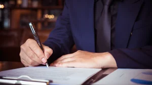 Un hombre con traje azul oscuro y corbata negra firma un documento con una pluma estilográfica sobre una mesa de madera. Sus manos están enfocadas mientras sostiene el bolígrafo con precisión. Junto al documento, se observan otros papeles con gráficos, lo que sugiere una situación formal de trabajo o negocios. La escena representa la firma de un contrato de agencia internacional, en un ambiente profesional y sobrio.