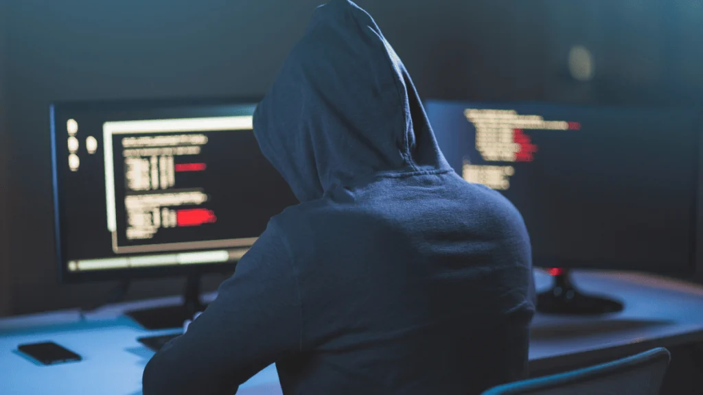 Persona vestida con una sudadera con capucha, vista de espaldas, sentada frente a dos monitores en una habitación con poca luz. Las pantallas muestran líneas de código con texto resaltado en rojo, lo que sugiere una actividad informática avanzada o potencialmente relacionada con la ciberseguridad o el hacking. La escena transmite un ambiente de anonimato y concentración.