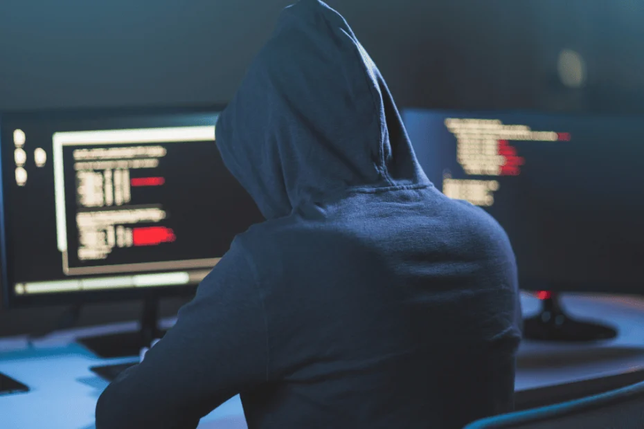 Persona vestida con una sudadera con capucha, vista de espaldas, sentada frente a dos monitores en una habitación con poca luz. Las pantallas muestran líneas de código con texto resaltado en rojo, lo que sugiere una actividad informática avanzada o potencialmente relacionada con la ciberseguridad o el hacking. La escena transmite un ambiente de anonimato y concentración.