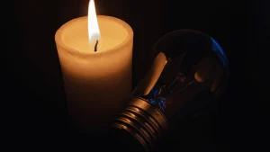 Una vela encendida ilumina suavemente una bombilla apagada, ambas colocadas sobre un fondo oscuro que sugiere una falta general de electricidad. La escena transmite una sensación de precariedad energética, evocando el contraste entre lo antiguo y lo moderno. Esta imagen sirve como metáfora visual del artículo titulado "Derechos laborales en España tras el gran apagón", representando el regreso a soluciones básicas en un contexto donde la tecnología y los servicios esenciales han fallado, y planteando la reflexión sobre cómo estos cortes afectan a los trabajadores y sus condiciones laborales.