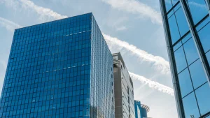 Fotografía tomada desde un ángulo bajo que muestra varios rascacielos modernos con fachadas de vidrio azul, reflejando el cielo despejado. Al fondo, trazos blancos de estelas aéreas cruzan diagonalmente el cielo. La imagen transmite dinamismo y expansión, evocando el mundo corporativo donde ocurren procesos estratégicos como las fusiones inversas.