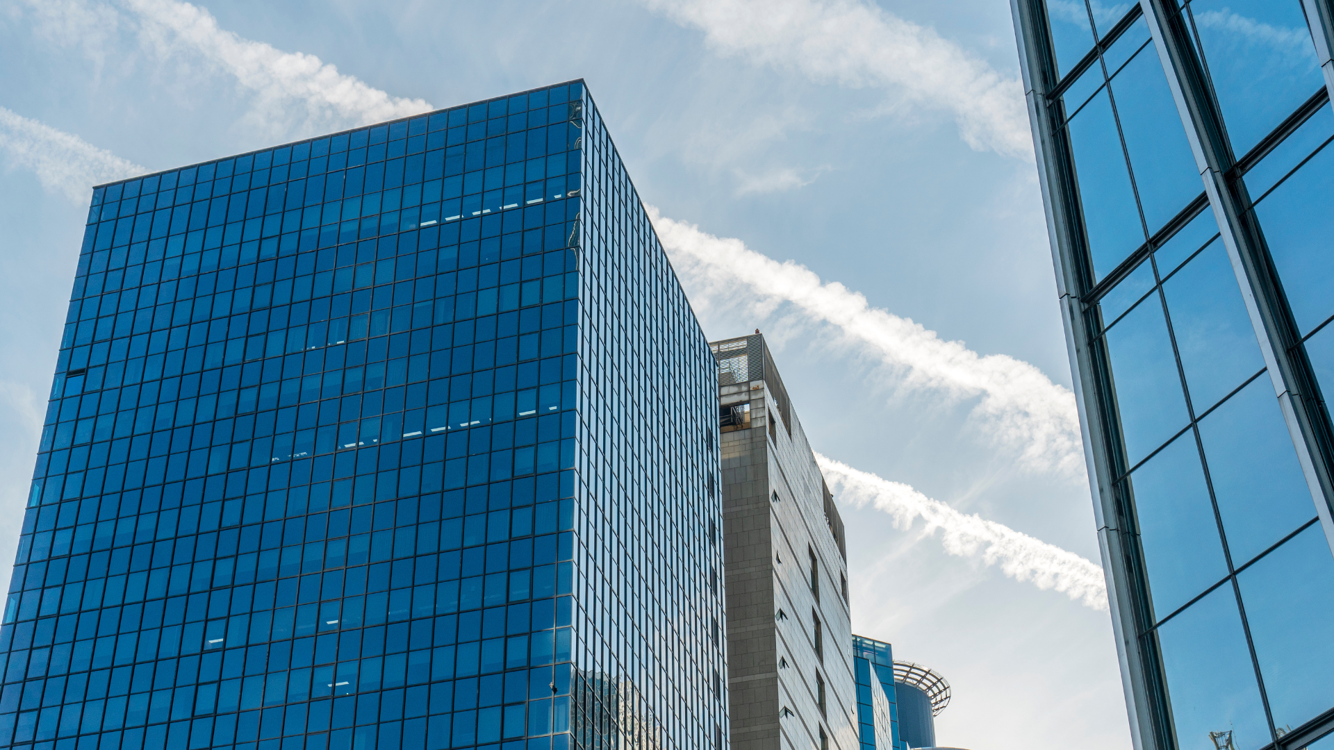 Fotografía tomada desde un ángulo bajo que muestra varios rascacielos modernos con fachadas de vidrio azul, reflejando el cielo despejado. Al fondo, trazos blancos de estelas aéreas cruzan diagonalmente el cielo. La imagen transmite dinamismo y expansión, evocando el mundo corporativo donde ocurren procesos estratégicos como las fusiones inversas.