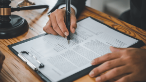 Primer plano de las manos de una persona con traje firmando un documento legal sobre una carpeta con sujetapapeles, sobre una mesa de madera. A la izquierda se observa un mazo de juez, lo que sugiere un contexto jurídico o notarial. La escena transmite formalidad y seriedad, común en procesos legales como contratos, testamentos o acuerdos.
