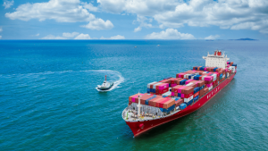 Un gran buque portacontenedores de color rojo, con la palabra "NAVIOS" visible en el casco, navega por un mar de aguas azules bajo un cielo parcialmente nublado. El barco transporta cientos de contenedores multicolores, organizados en filas sobre su cubierta. A su lado, una embarcación más pequeña, probablemente un barco piloto, lo acompaña haciendo un giro en el agua y dejando una estela blanca. En el horizonte se distinguen otras naves y una línea de islas lejanas. La imagen representa una escena típica del comercio marítimo y la logística internacional