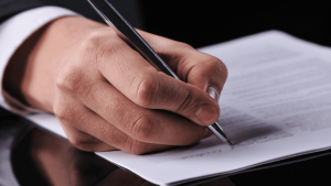 Mano de una persona con traje formal firmando un documento con bolígrafo negro sobre una mesa oscura. El encuadre enfoca en detalle el momento de la firma, donde se distingue texto impreso en el papel, aunque no es legible. La escena representa la formalización de una carta de intención, un paso clave en acuerdos comerciales, negociaciones laborales o procesos legales.