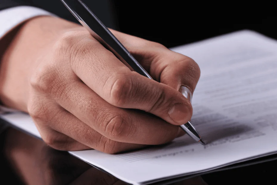 Mano de una persona con traje formal firmando un documento con bolígrafo negro sobre una mesa oscura. El encuadre enfoca en detalle el momento de la firma, donde se distingue texto impreso en el papel, aunque no es legible. La escena representa la formalización de una carta de intención, un paso clave en acuerdos comerciales, negociaciones laborales o procesos legales.