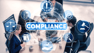 Imagen de un grupo de profesionales reunidos alrededor de una mesa de trabajo, vistos desde arriba, revisando documentos y usando dispositivos electrónicos. En el centro, se destaca la palabra “COMPLIANCE” (cumplimiento normativo) en letras grandes, rodeada por íconos digitales relacionados con el tema: una balanza de la justicia, un martillo de juez, una lista de verificación, engranajes con una marca de verificación, documentos legales, libros, una mano con una marca de aprobación y una lupa. La imagen transmite un entorno corporativo enfocado en la conformidad con normativas y estándares legales.