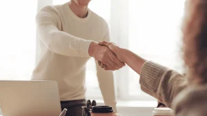 Dos personas se dan la mano en señal de acuerdo o bienvenida, en un entorno de oficina iluminado por luz natural. Una de ellas está de pie, con un suéter claro, mientras la otra permanece sentada, con un suéter de punto gris. En primer plano hay una computadora portátil, una libreta con bolígrafos y vasos de café desechables, sugiriendo un ambiente de reunión profesional o entrevista de trabajo.