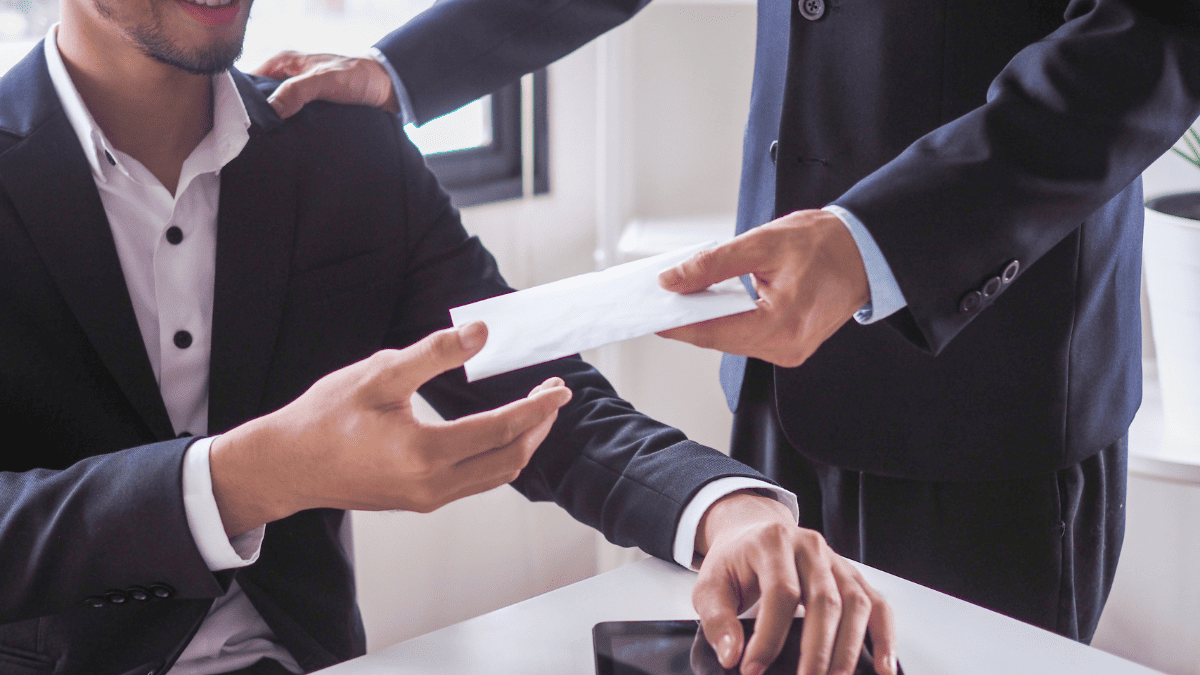 Dos hombres vestidos con traje formal interactúan en una oficina iluminada por luz natural. Uno de ellos, sentado, recibe un sobre blanco de manos del otro, que se mantiene de pie y le coloca la mano en el hombro en señal de apoyo. Sobre el escritorio hay una tableta. La escena representa el momento en que un particular recibe una indemnización
