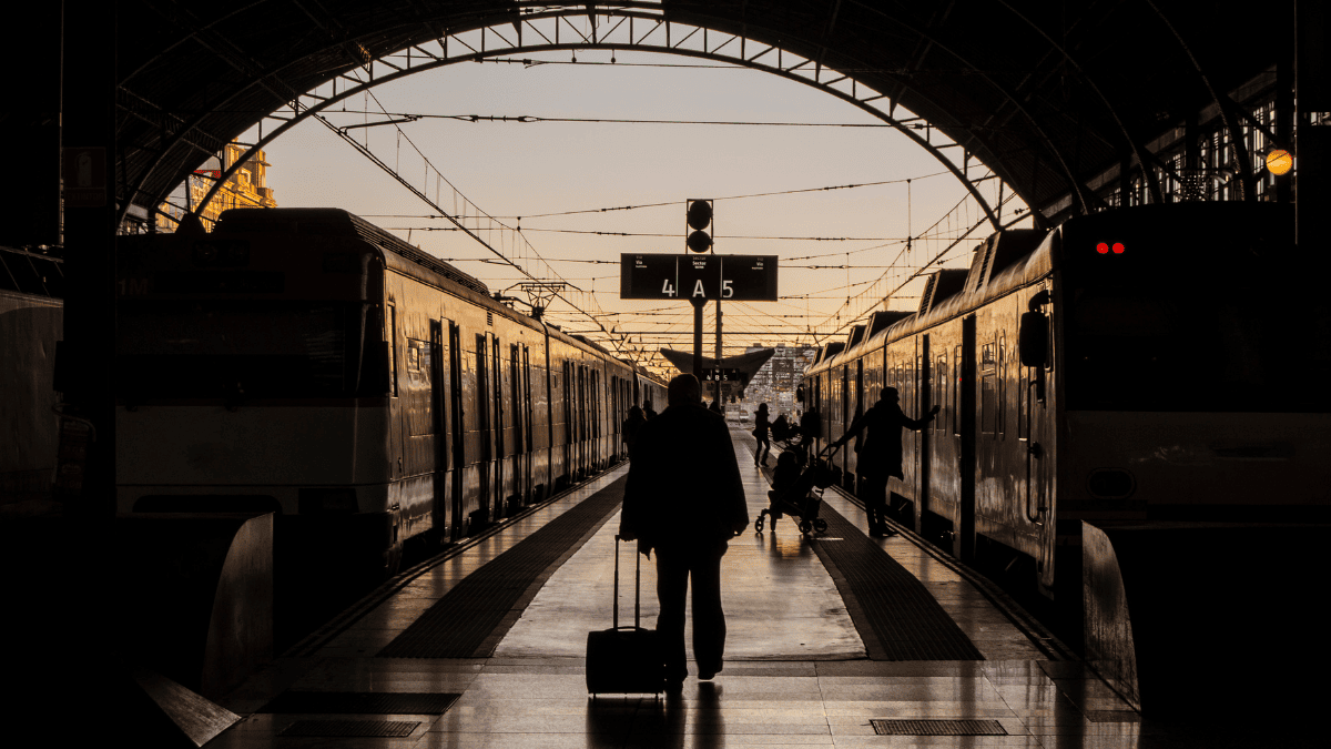 En una estación de tren iluminada por la luz dorada del atardecer, una persona camina por el andén central arrastrando una maleta con ruedas, mientras a ambos lados se encuentran dos trenes detenidos. La silueta del viajero está en primer plano, destacando contra el fondo claro. Más al fondo, se ven otras personas, incluyendo una figura empujando un coche de bebé y otra subiendo a un vagón. Sobre el andén cuelga un cartel que indica las vías 4A y 5.