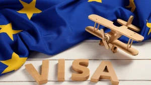 Sobre una superficie de madera blanca se encuentra una bandera de la Unión Europea arrugada, con fondo azul y estrellas amarillas. Delante de la bandera hay un avión de juguete de madera estilo biplano y, en primer plano, las letras de madera que forman la palabra "VISA". La imagen evoca el tema de los viajes a Europa y la obtención de visado Schengen.