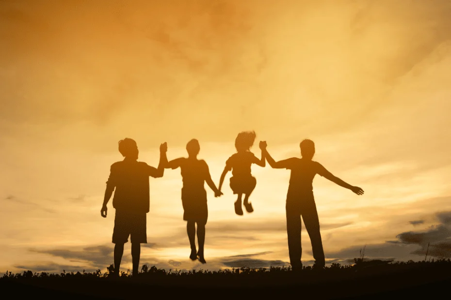 Silueta de una familia de cuatro personas al atardecer, con un cielo naranja y nubes suaves de fondo. Dos adultos y un niño están de pie, mientras una niña pequeña es levantada en el aire por las manos de los adultos a cada lado, captando un momento de alegría y unión familiar al aire libre. La escena transmite calidez, amor y convivencia familiar.