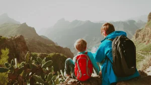 Un adulto y un niño, ambos con mochilas de senderismo, están sentados sobre una roca en un paisaje montañoso, mirando un mapa juntos. La escena transmite un momento de planificación durante una excursión en la naturaleza. Al fondo se aprecian montañas escarpadas cubiertas de vegetación y un cielo ligeramente brumoso. La imagen sugiere un viaje compartido, ideal para ilustrar temas relacionados con viajes con hijos menores y custodia compartida.