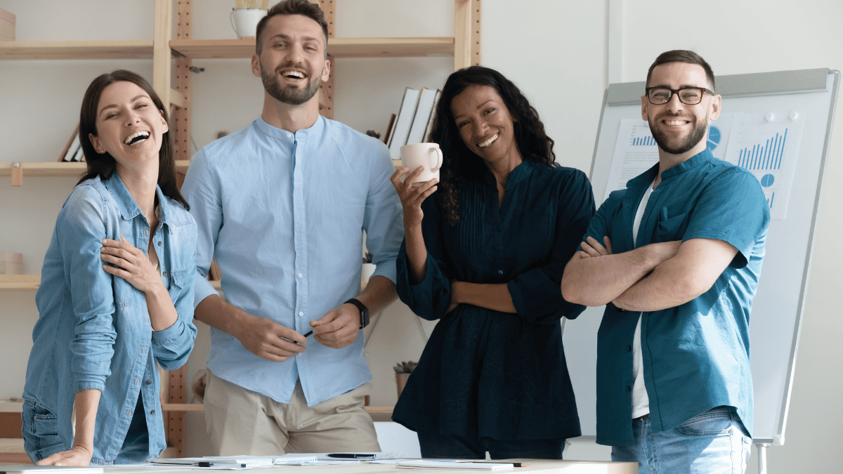 Grupo de cuatro personas jóvenes en una oficina, sonriendo y mostrando una actitud relajada y amigable. Están de pie frente a una mesa con documentos y una pizarra con gráficos en el fondo, lo que sugiere un ambiente laboral o reunión de trabajo. Dos hombres y dos mujeres, vestidos de manera informal, representan diversidad y colaboración en un entorno profesional moderno.