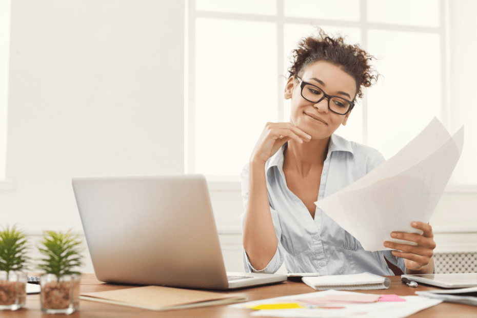 Mujer joven con gafas, camisa azul clara y cabello rizado recogido, sentada frente a un ordenador portátil mientras revisa papeles en una oficina con luz natural.