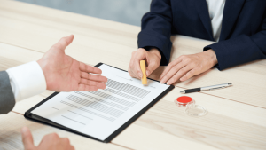 Dos personas sentadas frente a frente revisan y firman un contrato de trabajo sobre una mesa, con un sello y un bolígrafo al lado.