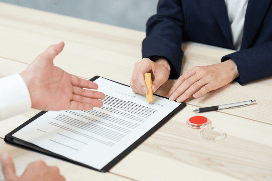 Dos personas sentadas frente a frente revisan y firman un contrato de trabajo sobre una mesa, con un sello y un bolígrafo al lado.
