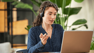 Mujer joven con auriculares y micrófono atendiendo una videollamada profesional desde su portátil, ejemplo de teletrabajo internacional en casa.